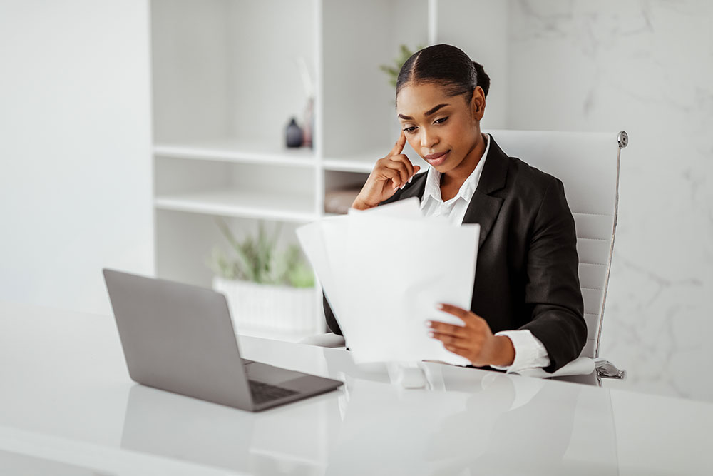 Business paperwork. Black businesswoman reading papers, sitting in front of laptop computer in light modern office. Female manager looking through documentation, bills and correspondence