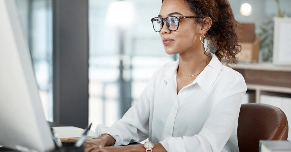 A woman with glasses and a white button-down is writing a professional email on her computer. The background is blurred but she is in an office with plenty of natural light.