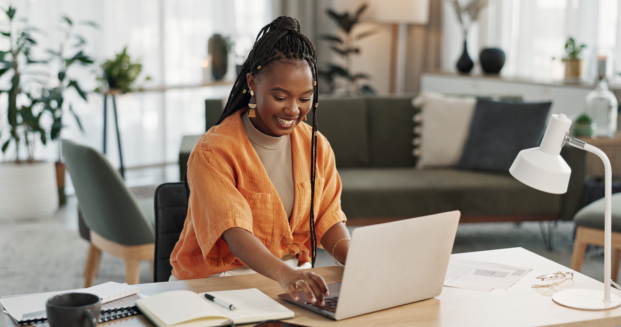 A black woman with long hair is at a desk writing a professional email. While the background is blurred, there is living room furniture behind her and a lot of light coming through the windows. The woman is smiling and is wearing an orange blouse.