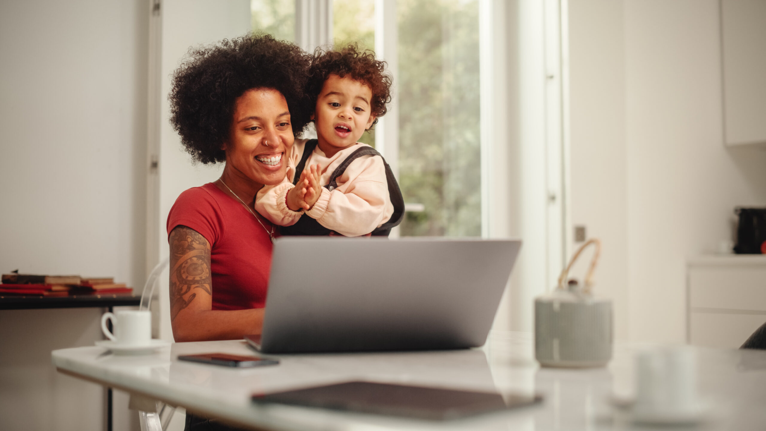 A black woman with a tattoo is at her kitchen table with a toddler in her arms while she writes a professional email. She and her child are smiling and the kitchen is light and bright.